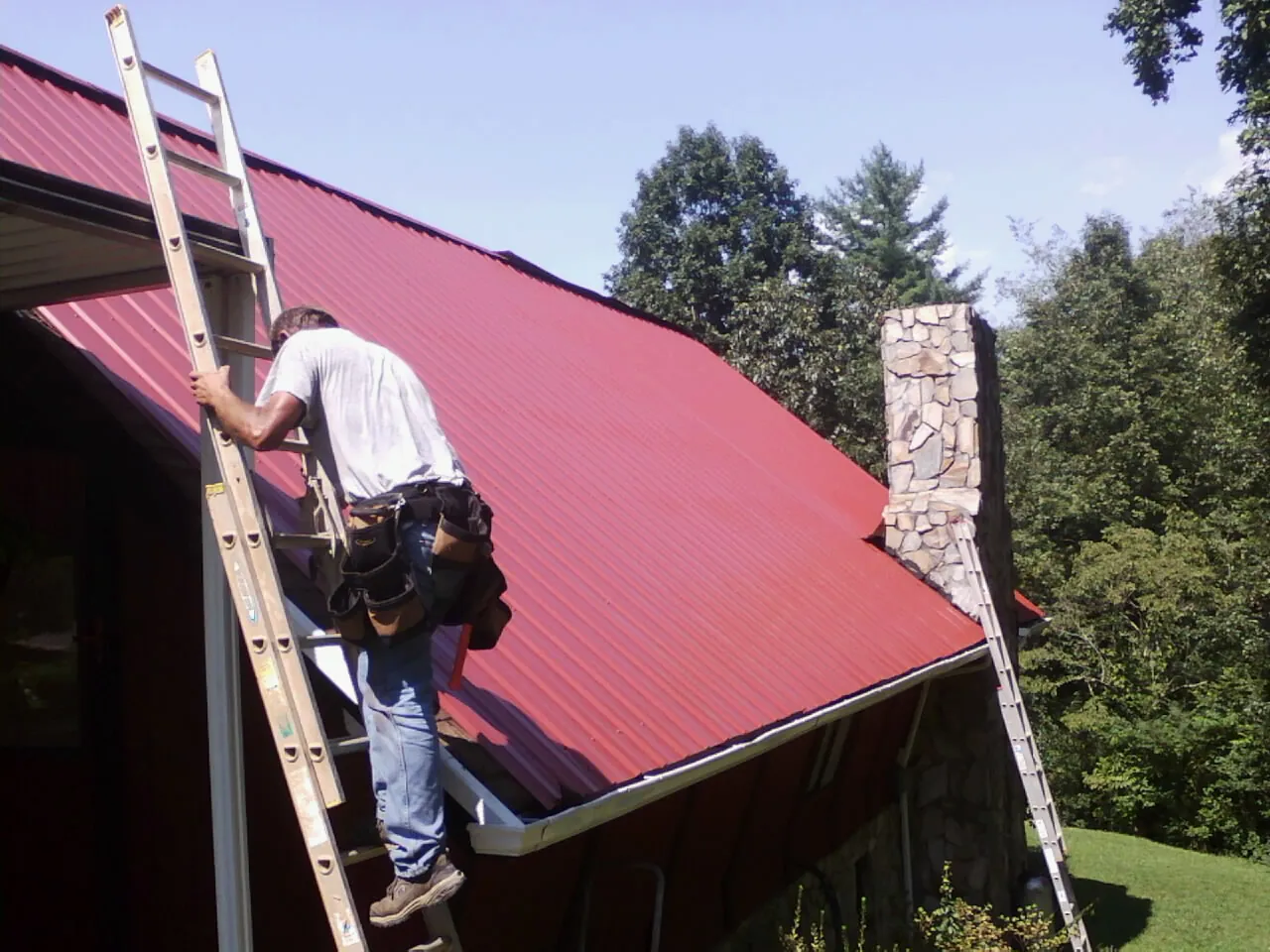 Roofer on a ladder beside a red metal roof and stone chimney — Soffit & Fascia Repair by Roofing Brothers in Onset