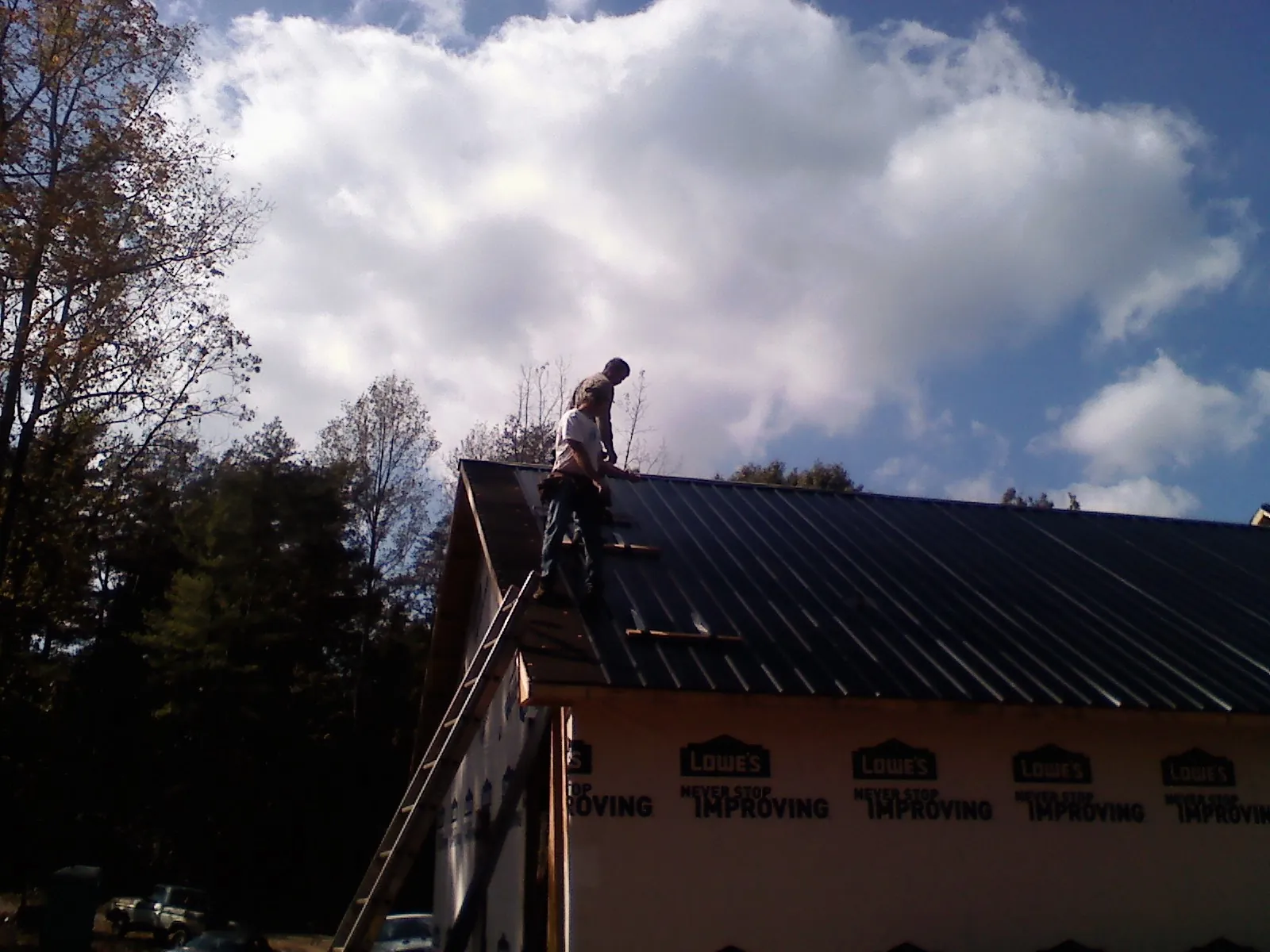 Roofer fastening dark metal panels high against a cloudy sky, from a Roof Inspection job in Onset